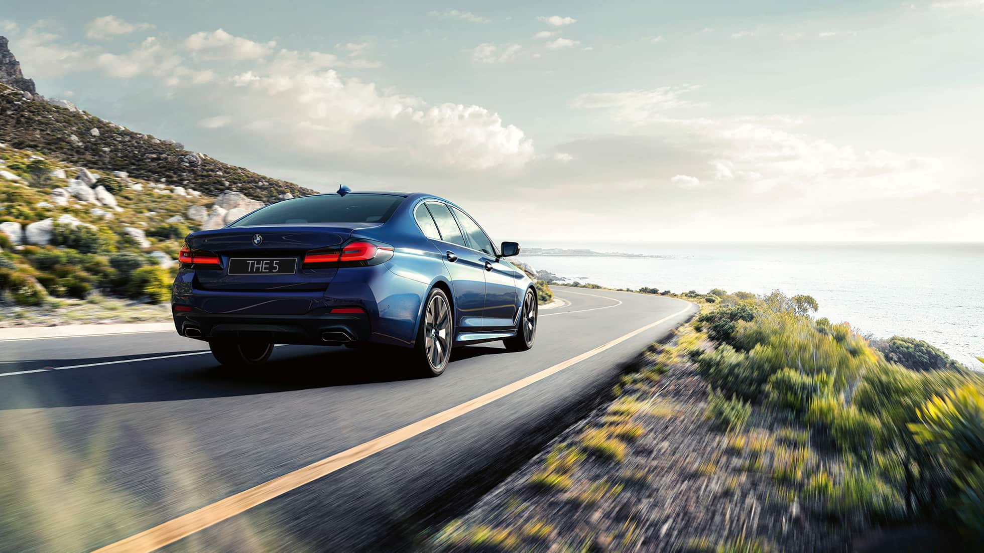 Динамичный BMW 5 серии G30, цвет «Синий Фитоник», вид сзади с поворотом в три четверти, в движении